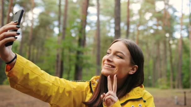 Retrato de la linda joven haciendo selfies en el teléfono inteligente, sonriendo y mostrando el gesto de victoria en el bosque. De cerca. Al aire libre — Vídeo de stock