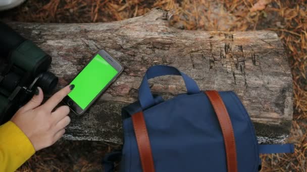 Vista dall'alto su donne che scorrono a mano sullo smart phone nero che si trova sul tronco di legno nella foresta vicina con binocolo e zaino. Schermo verde. Chiave cromatica. All'aperto. Da vicino. — Video Stock