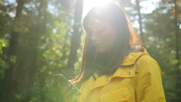 Gros plan de la jeune belle femme en manteau jaune tapant sur son téléphone intelligent au milieu de la forêt sous la lumière. Portrait. À l'extérieur — Video