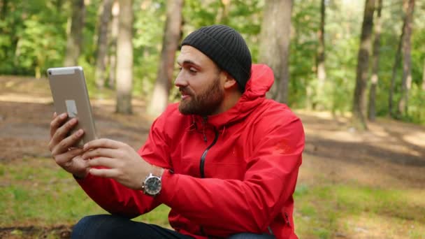 Retrato del joven guapo que tiene un video chat en su tableta en el hermoso bosque y muestra el bosque a la cámara. Exterior — Vídeos de Stock