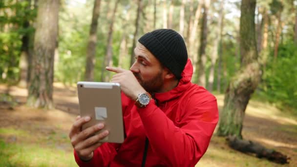 Retrato del joven senderista guapo chateando en su tableta en el hermoso bosque soleado y mostrando el bosque a la cámara. Al aire libre — Vídeos de Stock