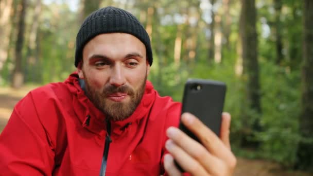 Primo piano del giovane bel video di escursionisti maschi che chiacchierano sullo smartphone nero, agitando la mano e mostrando il segno della vittoria nella bella foresta. Ritratto. All'aperto — Video Stock