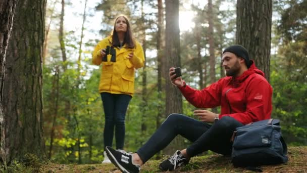 Pareja joven descansando en el bosque. Una mujer guapa mirando los prismáticos y un hombre haciendo selfies. Día soleado. Exterior — Vídeos de Stock