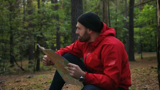 Atractivo joven con abrigo rojo y sombrero sentado en el bosque y leyendo un mapa. Retrato. Me pierdo. Exterior — Vídeos de Stock