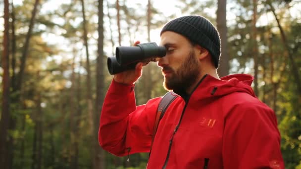 Porträtt av unga stiliga mannen med skägg står mitt i skogen och tittar i kikaren. Solig dag. Närbild. Utomhus — Stockvideo