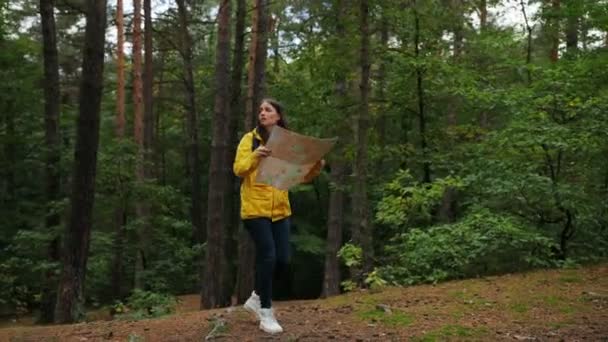 Молодая привлекательная женщина в желтом пальто и с рюкзаком потерялась в лесу. Испуганная женщина ищет выход с помощью карты. На открытом воздухе — стоковое видео