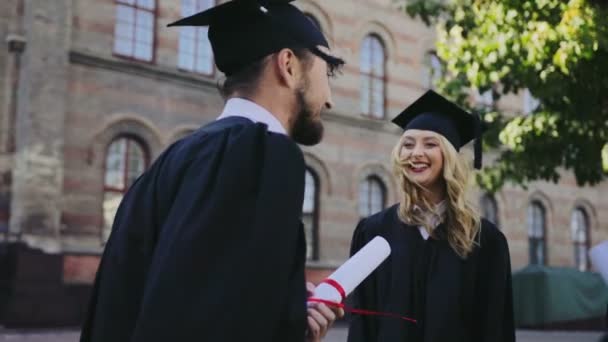 Hombres y mujeres jóvenes graduados hablando y riendo con ropa tradicional y gorras en su ceremonia de graduación. Al aire libre — Vídeo de stock
