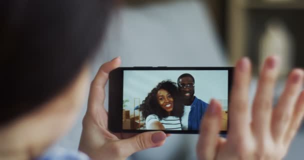 View over the shoulder on the young woman holding a smartphone on which having a web cam chat with African American young couple who moving in a new apartment and showing keys to the camera. Close up. — Stock Video