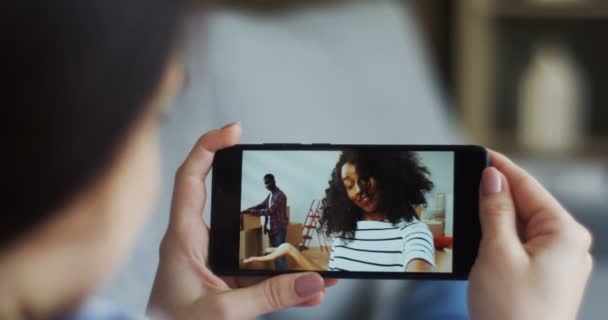 Traseira da jovem segurando um dispositivo de smartphone no qual ter uma webcam bate-papo com a jovem afro-americana que chamando seu namorado enquanto se mudam juntos em um novo apartamento. Fechar . — Vídeo de Stock