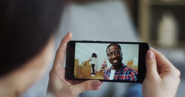 Baksidan av den unga kvinna som håller en smartphone enhet som har en web cam chatt med African American ung man vem ringer hans flickvän medan du flyttar ihop i en ny lägenhet. Närbild. — Stockvideo