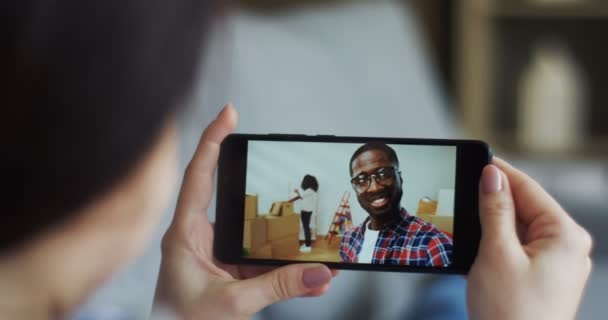 Hinten hält die junge Frau ein Smartphone in der Hand, auf dem sie mit einem afrikanisch-amerikanischen jungen Mann chatten kann, der seine Freundin anruft, während sie in eine neue Wohnung zieht. Nahaufnahme. — Stockvideo