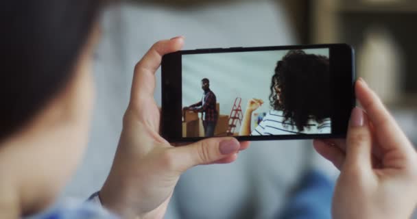 Visa över axeln på kaukasiska kvinnan med en svart smartphone som att ha en videochat med African American ung kvinna och hennes make medan de rör sig i ett nytt hus. Närbild. — Stockvideo