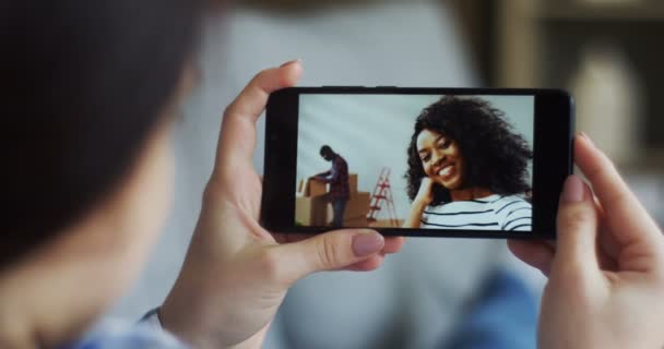 Nahaufnahme der brünetten Frau, die mit ihrer afrikanisch-amerikanischen Freundin, die mit ihrem Freund eine neue Wohnung bezieht, einen Videochat auf der Webcam des Smartphones führt. hinten. — Stockvideo