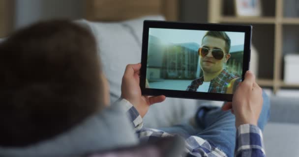 Крупный план черного планшетного устройства в руках человека, который разговаривает через веб-камеру с другом-мужчиной, лежащим на диване . — стоковое видео