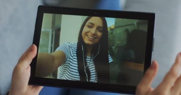 Tablette noire avec jeune femme charmante dans les écouteurs ayant un chat webcam sur son écran. Gros plan des mains féminines tenant un dispositif . — Video