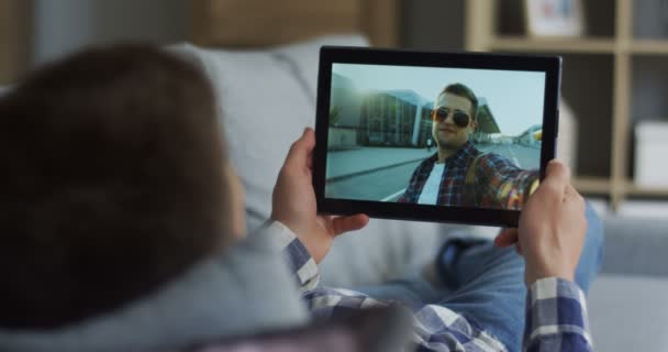 Вид через плечо на молодого человека, лежащего на диване и ведущего веб-камеру чата со своим другом, стоящим на улице на черном планшетном компьютере. Закрыть . — стоковое видео