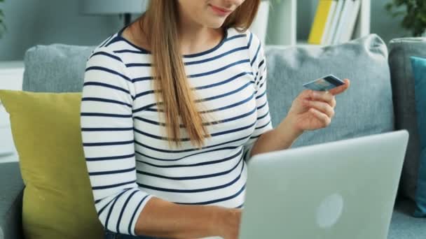 Retrato de la mujer bonita comprando en línea en el ordenador portátil e introduciendo los datos de la tarjeta de crédito mientras está sentado en el sofá en casa. En interiores — Vídeos de Stock