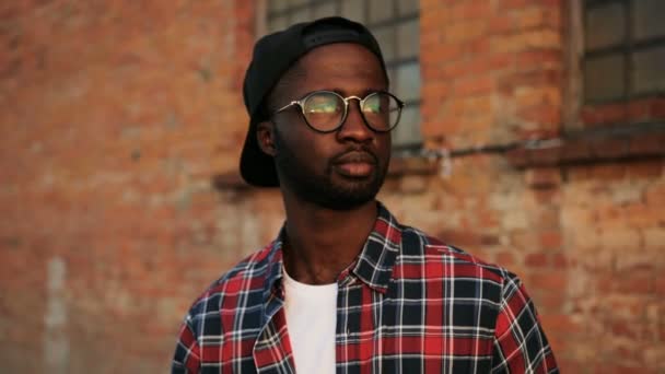 Close up de jovem afro-americano em sunglasess sorrindo para a câmera. O fundo da parede de tijolos. Espaço urbano. Lá fora. Retrato — Vídeo de Stock