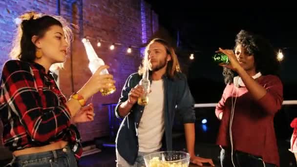 De jeunes amis multi-ethniques debout et buvant de la bière autour des tables à la fête sur le toit la nuit. Le fond du mur de briques. À l'extérieur — Video