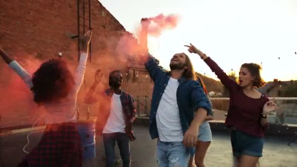 Jóvenes amigos de carreras mixtas se divierten con una granada de humo rosa en la fiesta en la azotea en el espacio urbano. El fondo del atardecer. Afuera. . — Vídeos de Stock