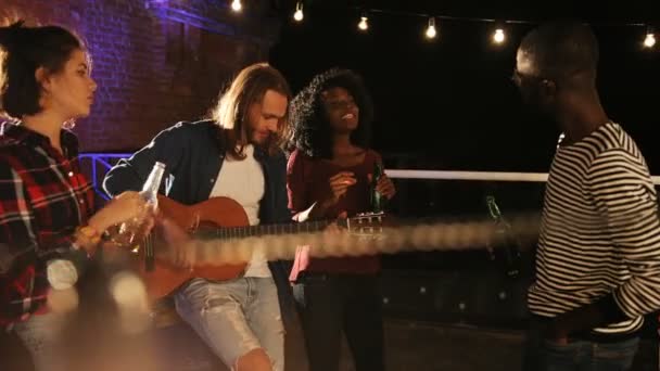 Attractive young people partying, singing and dancing while guy playing on the guitar on the rooftop party. At night. The brick wall with lights background. Outside. Multiethnic — Stock Video