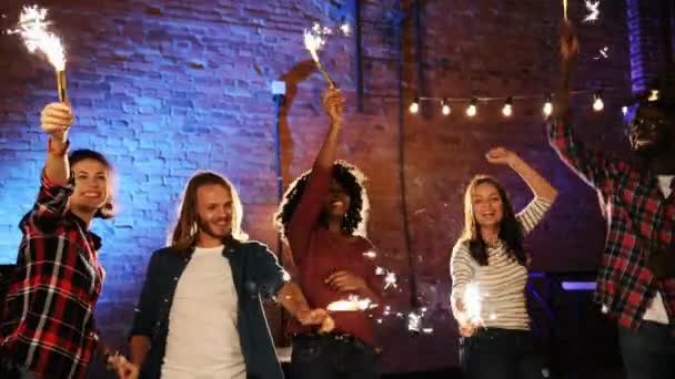 Young friends dancing and playing with golden sparkler sticks at the rooftop party at night. The brick wall with lights background. Outdoor. Mixed races — Stock Video