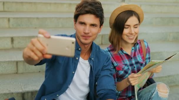 Trendiga unga paret i stan sitter på trappan tar en selfie utomhus, hålla en stadskarta. — Stockvideo
