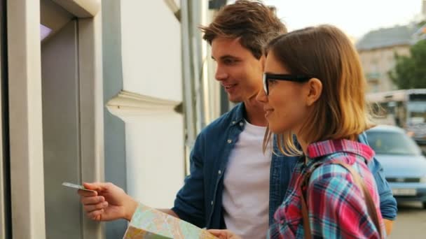 Touristic couple looking happily at each other in front of the ATM machine. Attractive caucasian woman is holding a city map. Outdoors. — Stock Video