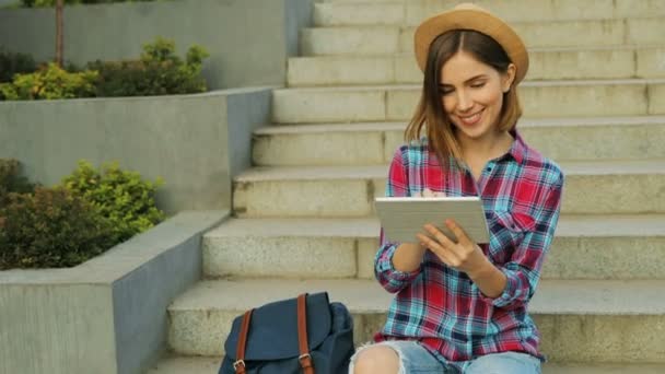 Молода стильна дівчина сидить на сходах і тримається за руки за планшет. Студент спілкується в соціальних мережах з друзями, вводячи повідомлення зі сходами на задньому плані . — стокове відео