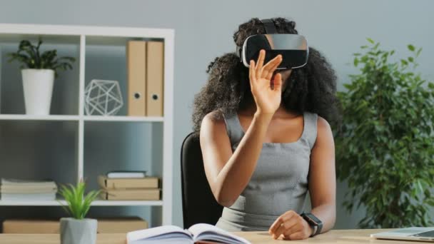 Jovem afro-americana muito crespos usando fone de ouvido realidade virtual, óculos VR enquanto se senta na mesa no escritório. Interior — Vídeo de Stock