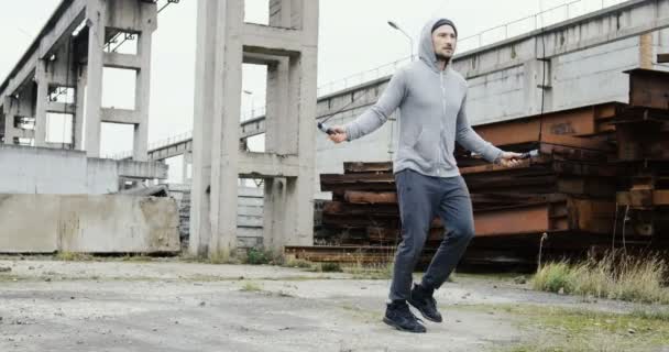 Ein kaukasischer Sportler in grauer Trainingskleidung mit Kapuze springt im nebelverhangenen Morgen auf dem Springseil in den Ruinen alter Industrieanlagen. im Freien — Stockvideo