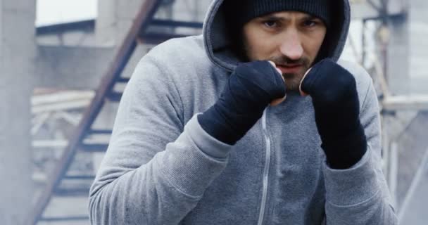 Retrato do jovem boxeador batendo na frente da câmera em um capuz cinza e com as mãos embrulhadas no dia cinzento nublado frio. Instalações industriais abandonadas. Ao ar livre — Vídeo de Stock