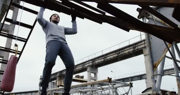 Joven deportista guapo que se detiene en la construcción de metal en las ruinas de viejas instalaciones industriales abandonadas en el día de niebla fría. Exterior — Vídeo de stock