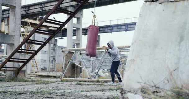 Lejos del joven boxeador guapo con barba pateando una bolsa roja en el distrito industrial abandonado en la mañana brumosa de Cols. Al aire libre — Vídeos de Stock