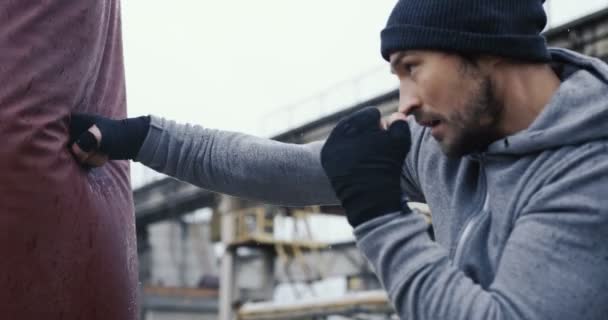 Närbild på den manliga boxare att göra omfattande sparkar på röda boxning påsen utomhus på den kalla dimmiga dagen. Övergivna anläggningen territorium. — Stockvideo