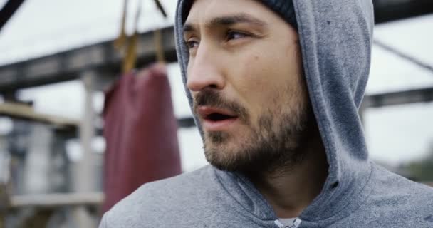 Gros plan sur le sportif fatigué attrayant dans une capuche respirant à peine après l'entraînement de boxe sur l'air frais. Installations industrielles ruines. En plein air. Portrait — Video