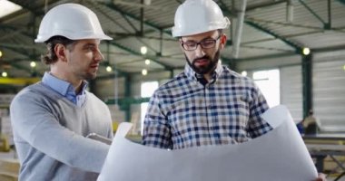 Miğferli, beyaz, yakışıklı iki adam büyük kağıda çizimlerle bakıp bunu tartışıyor. Fabrika alanı. Kapalı alanda. Portre. Kapat.