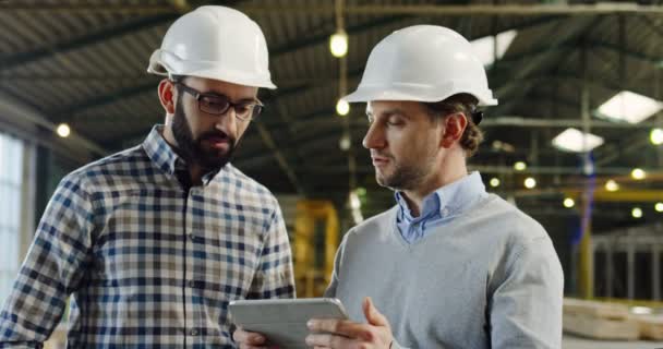 Two Caucasian men in helmets talking and looking at the tablet device screen, than rising their looks into the camera and smiling. The empty factory space background. Indoor. Portrait. Close up — Stock Video