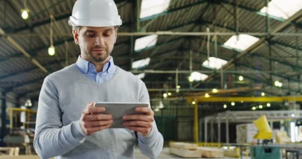 Ritratto di un bell'uomo ingegnere in un casco che registra e scorre sul suo tablet e si guarda intorno nel grande magazzino della fabbrica. Interni . — Video Stock