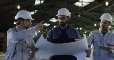 Kask ile büyük üç mühendislerince yürüyüş ve onların proje tartışıyor çizimler kağıt. İçinde büyük bir fabrikada planlama.