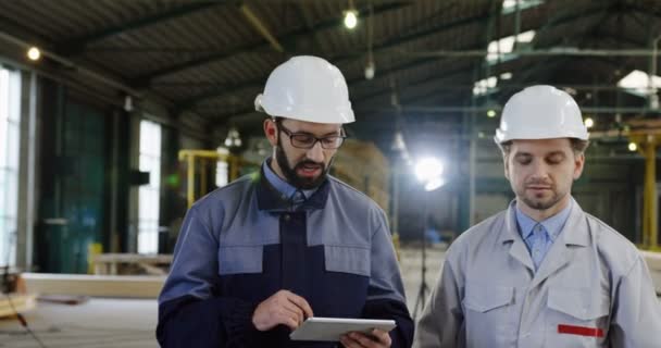 两名戴头盔的男性工程师拿着平板设备走在一家大型家具厂内讨论新项目。工作过程的背景。人像镜头 — 图库视频影像