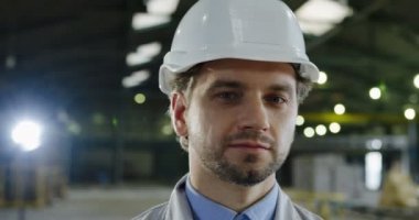 Kafası kameraya dönük ve gülümseyen kasklı çekici beyaz mühendisin yakın çekimi. Bulanık arka plandaki büyük fabrika evi. Portre çekimi