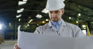 Çekici bir beyaz adam, mühendis çizimleri ile büyük kağıt onun elinde tutan ve dikkatle bakarak bir kask içinde portresi. Arka planda büyük fabrika. Yakın çekim