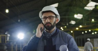 Kafkaslı beyaz fabrika işçisi büyük sanayi odasının ortasında akıllı telefonla konuşuyor. Kapalı alanda. Portre. Kapat.