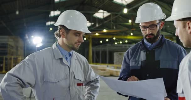 Primer plano de ingenieros en cascos con grandes dibujos en papel trabajando y planificando el próximo proyecto. Dentro de una gran planta. Retrato plano — Vídeos de Stock