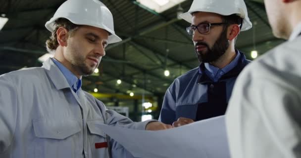 戴着头盔的男性工程师拿着大纸图纸谈论、讨论和规划下一个工作项目。在一个大工厂里肖像。特写 — 图库视频影像