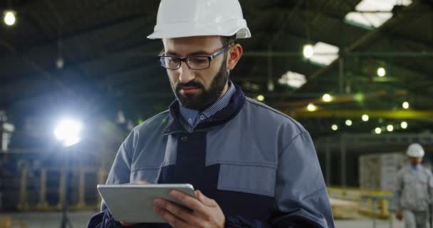 Gutaussehender Fabrikarbeiter mit Helm, der inmitten großer Industrieflächen am Tablet-Computer arbeitet. drinnen. Porträtaufnahme. Nahaufnahme — Stockvideo