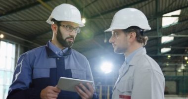 Kasklı, tabletli iki fabrika işçisinin fotoğraf çekimi. Konuşuyorlar ve kameraya poz veriyorlar. Büyük bir fabrikanın içinde..