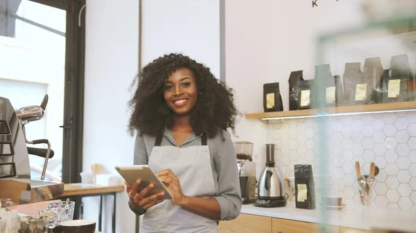 Πορτρέτο του θηλυκό αφρικανός barista με καφέ σγουρά μαλλιά χρησιμοποιώντας το tablet, κοιτάζοντας προς την κάμερα και χαμογελώντας στο καφέ καφέ. Πλευρική άποψη. Εικόνα Αρχείου