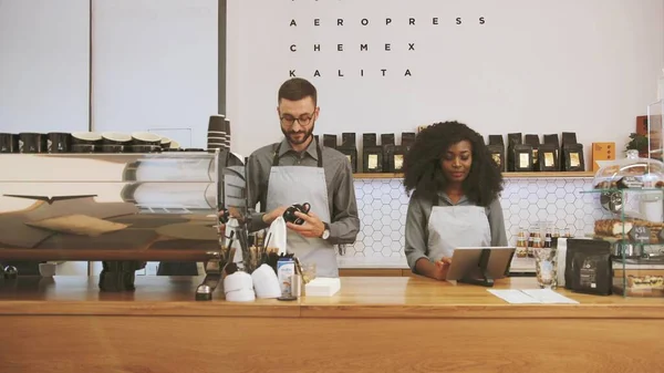 Hipster caucasian barista with beard and african young woman working in modern trendy coffee shop cafe, man washing cup and woman using laptop. Concept Preparing in for opening Small Business and Stock Photo